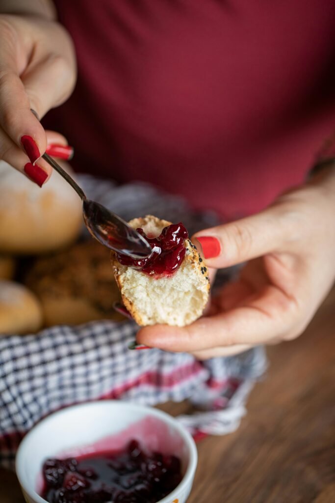 Close-up of a hand spreading berry jam on a freshly baked bread bun, highlighting homemade food comfort.